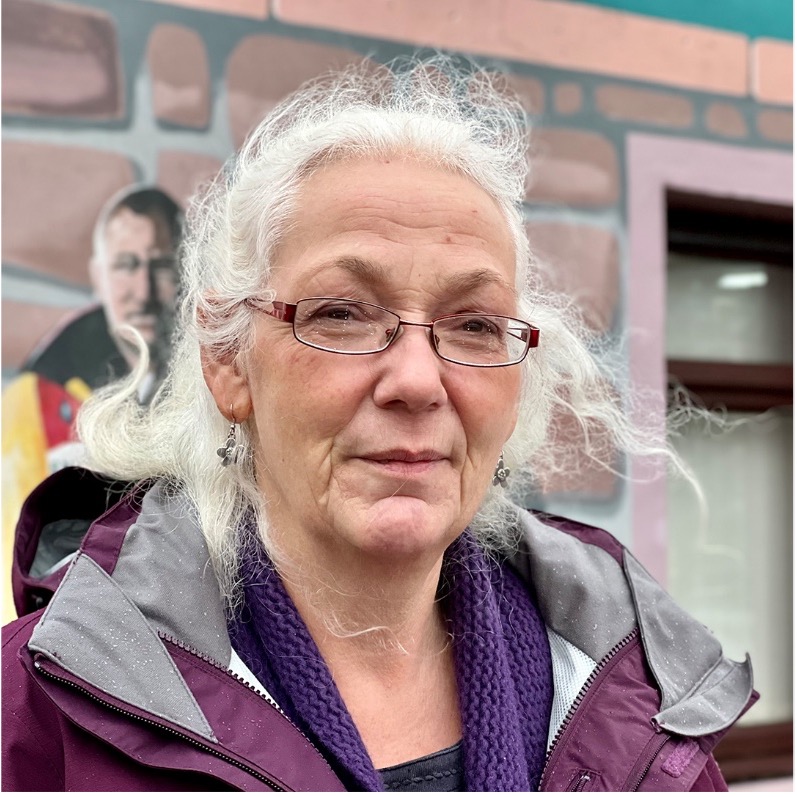 A portrait of Helen Ross. She is wearing glasses, a purple coat and has white hair. She is standing in front of a brick wall.