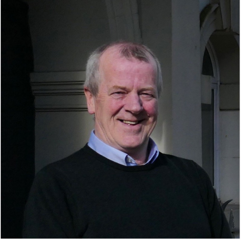 A portrait photo of Eric Spence. He is smiling, standing in front of a dark background, wearing a navy jumper. 