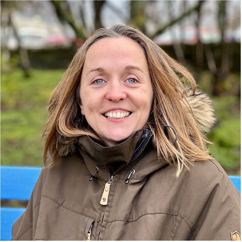 A portrait photo of Emma Whitham. She's sitting on a blue bench and wearing a brown jacket.