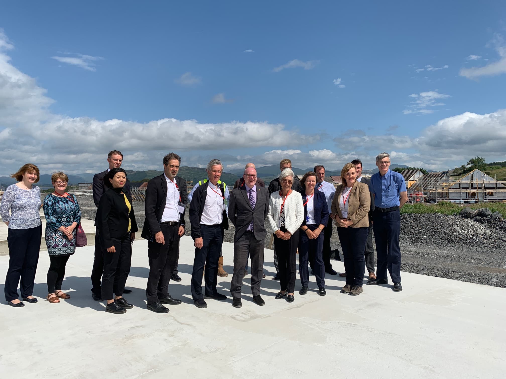 A group of people stand on a concrete foundation on a building site with the sea, hills and blue skies visible behind