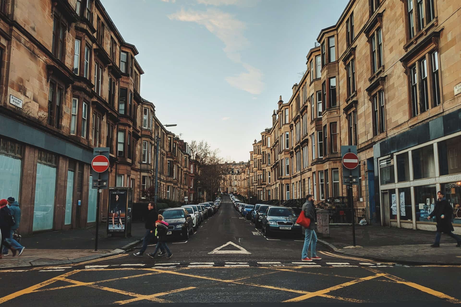 People walking across a road in Glasgow in front of a row of houses and cars.