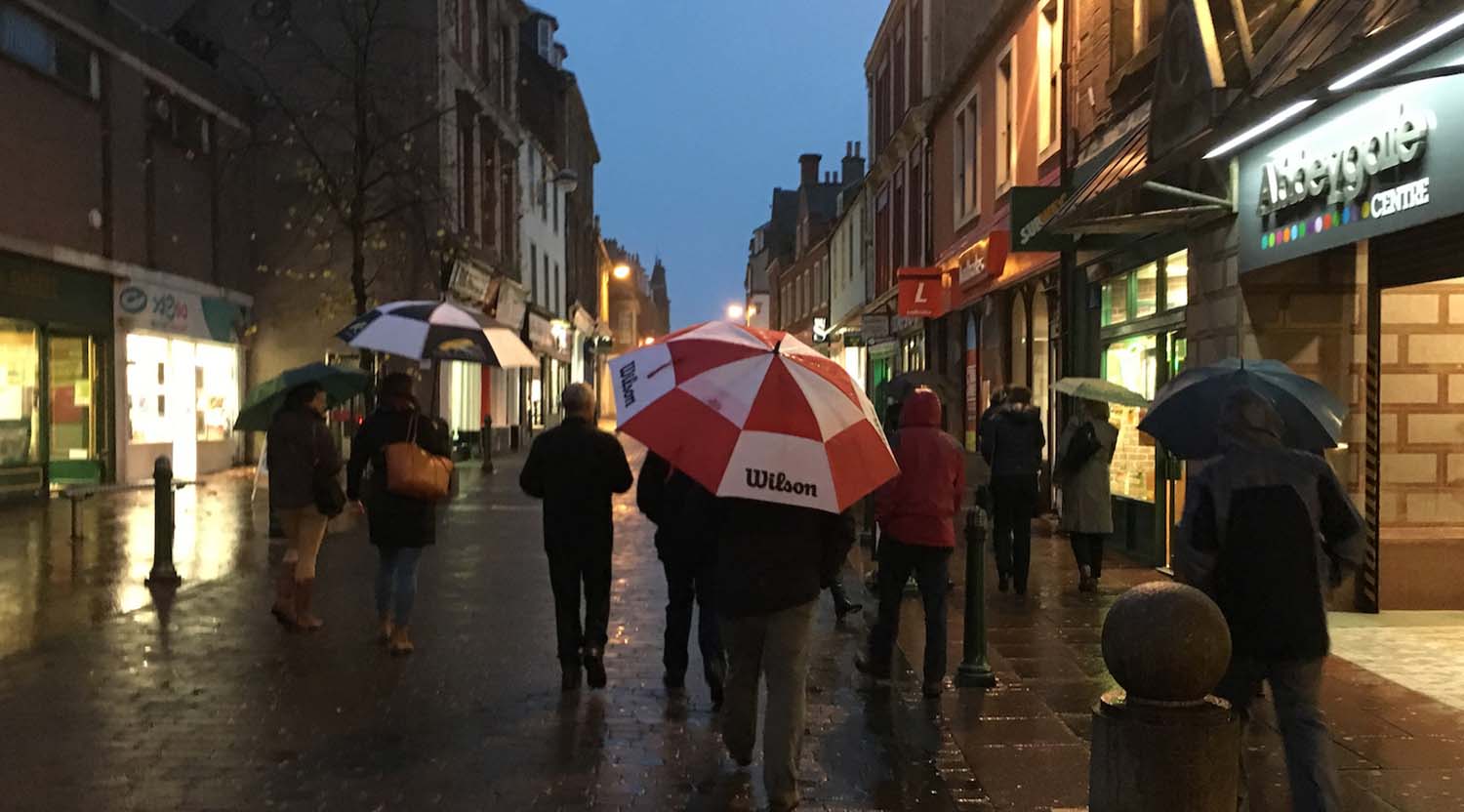 A group of people walking away from the camera at dusk through a high street