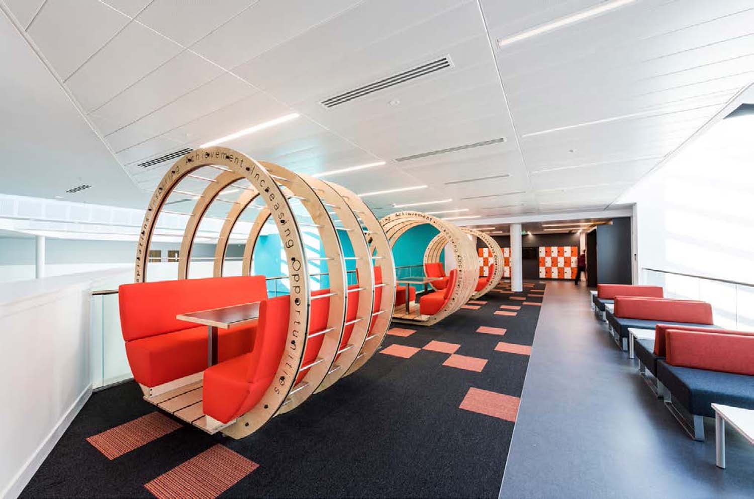 A circular wooden structure with orange upholstered seating inside set in a room with colourful seats