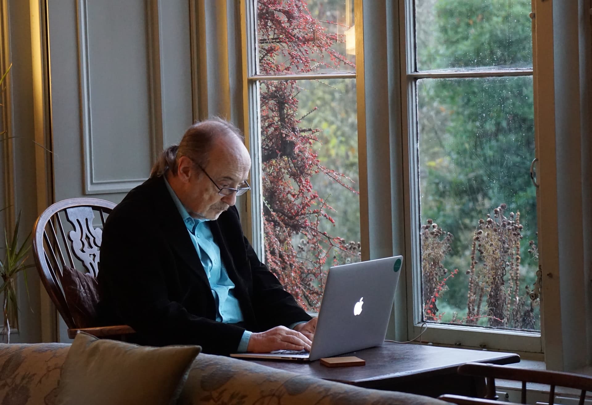 An elderly man using a laptop on a desk beside a window.