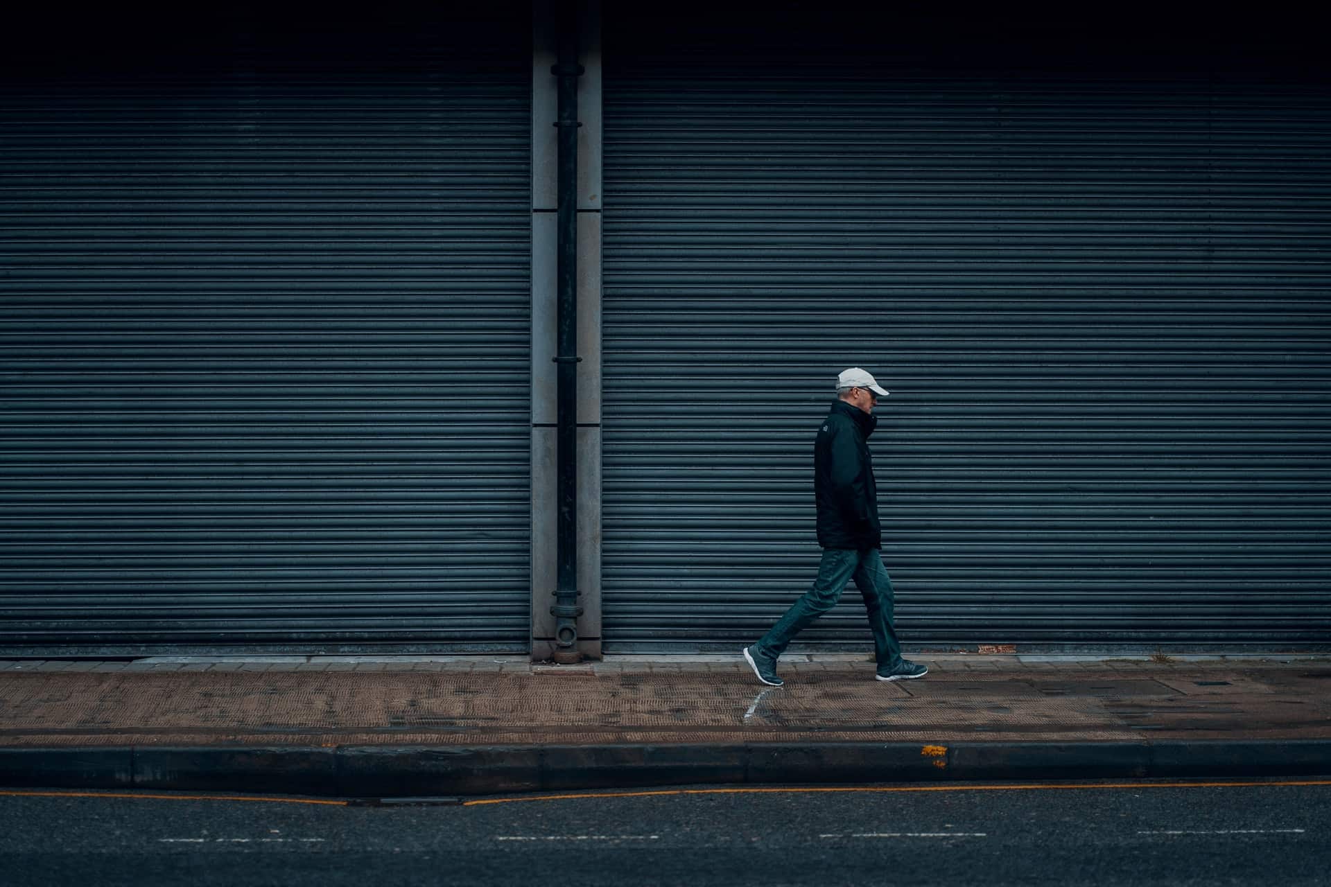 A man wearing a white hat and black jacket walking on a street pass a wall of shutters.