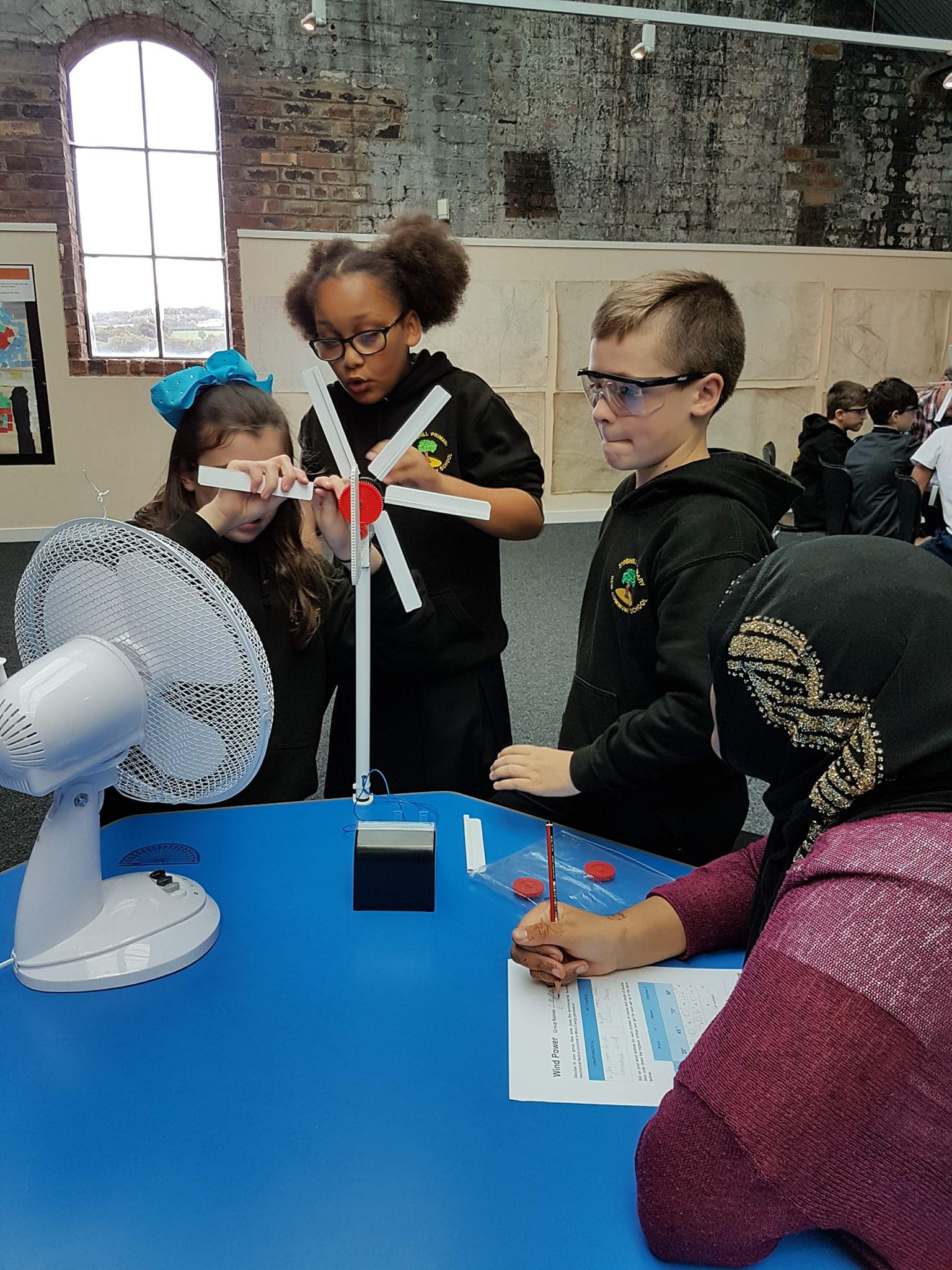 Four children are around a table building a model wind turbine in front of a fan.