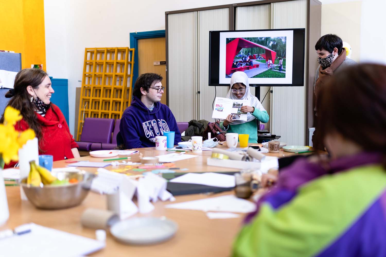 A young girl in a hijab presenting her work in a workshop to a group of people. A red canopy in the park is presented on the screen behind her.