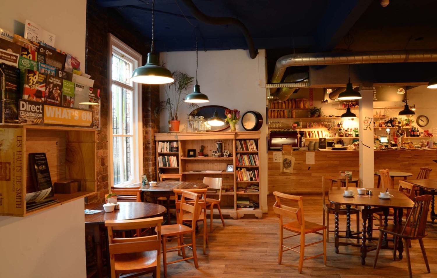 The Glad Cafe seating area with wooden tables and chairs, a bookshelf filled with books and dark green light pendants.