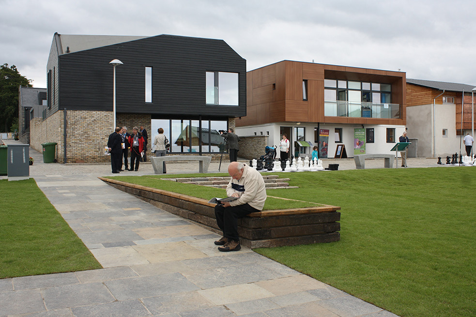 Two homes look over a village green on an overcast day. People are milling about the edge of the green and an older person in the foreground is sitting down reading.