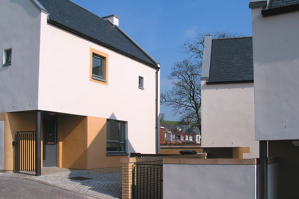 A close-up shot of a few homes with white walls and dark grey roofs against a bright blue sky. A leafless tree and a collection of houses are pictured in the background.