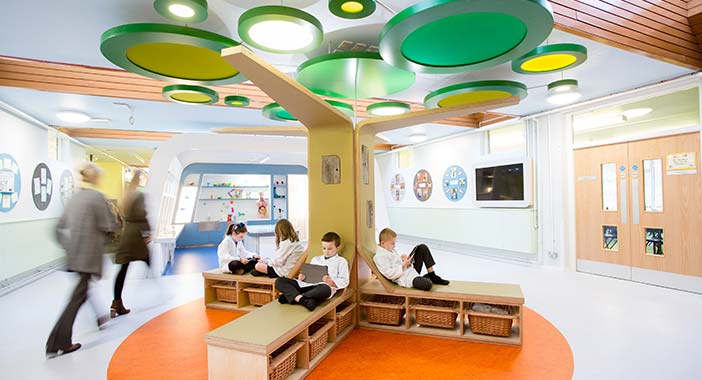 A wooden tree with seating underneath in the middle of a circulation area in a school with pupils walking through the space