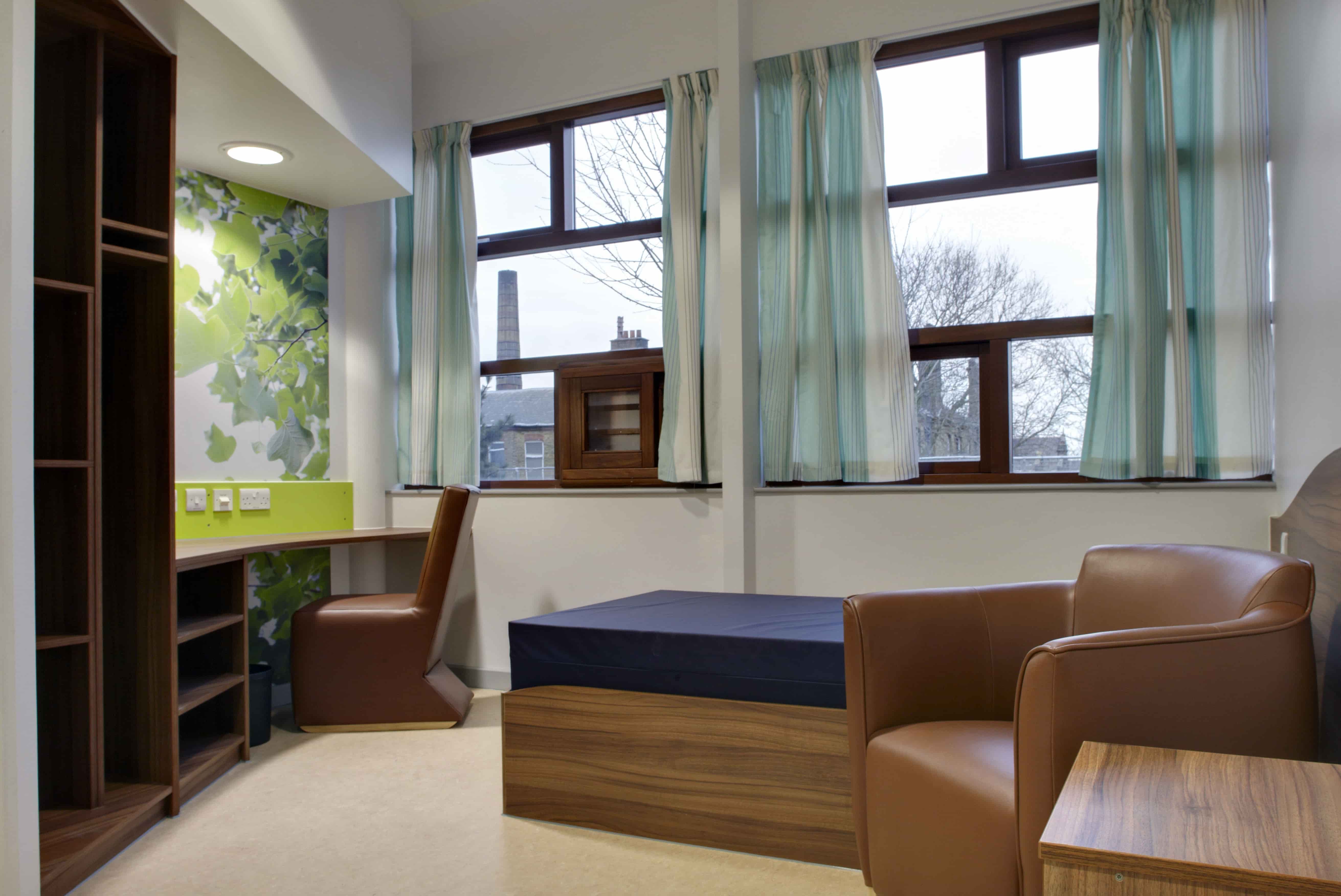 A single bedroom in Springfield University Hospital with a leather armchair beside it. A study desk and storage is positioned opposite the bed.