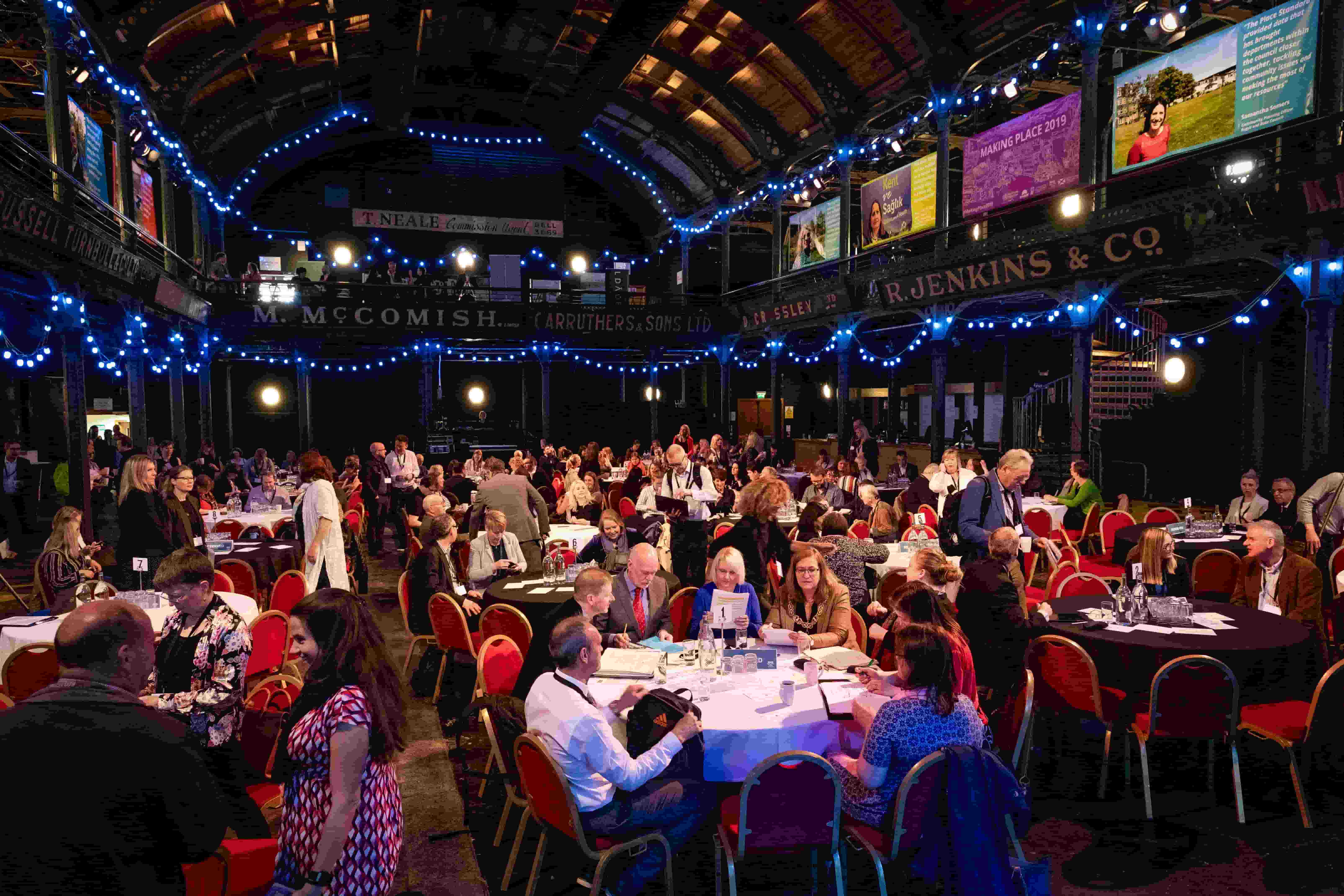 A conference taking place in a large room where the delegates are sitting around round tables with white table cloths and coloured exhibition panels hang from the balcony above