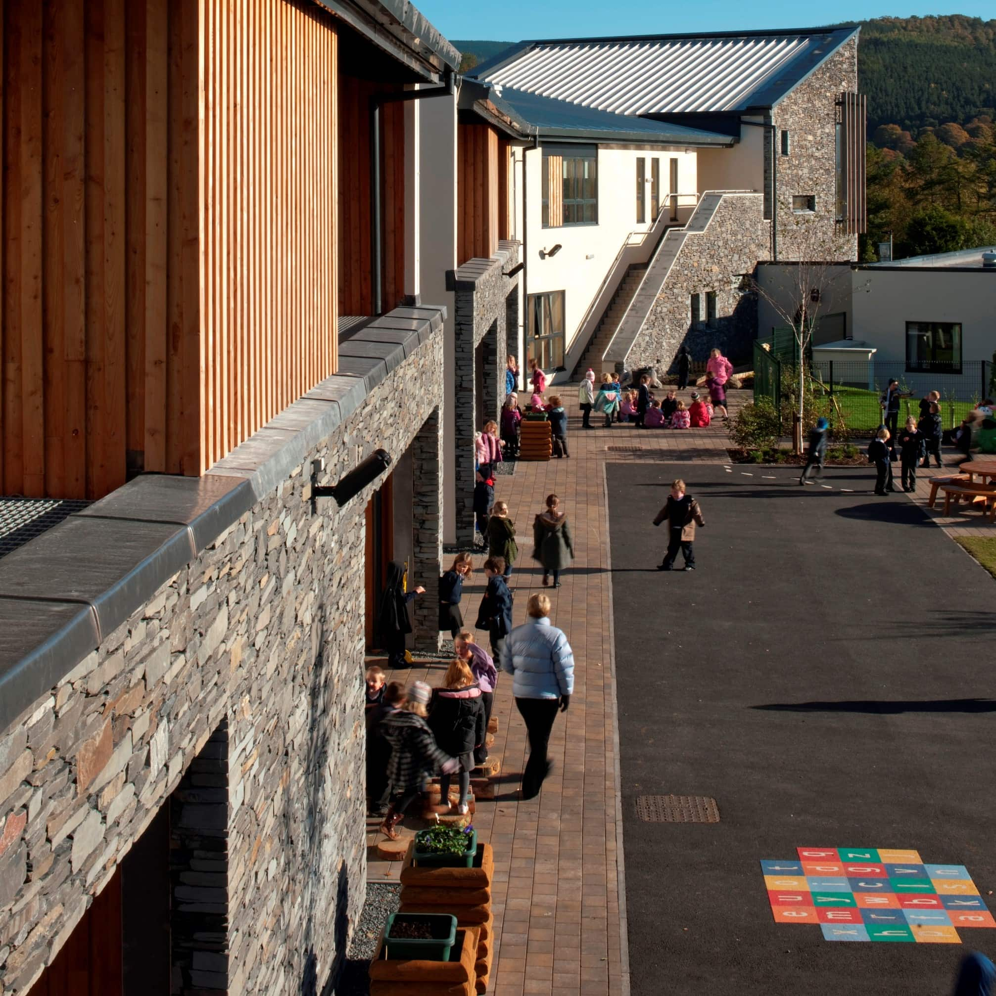 Looking down towards the ground floor exterior of Kingsland Primary School. Pupils and teachers are walking on the pavement next to the stone and wood façade building.