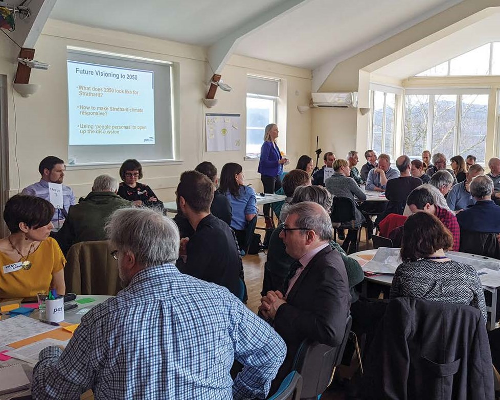 Workshop participants sitting at round tables. There is a presentation projected on the wall.