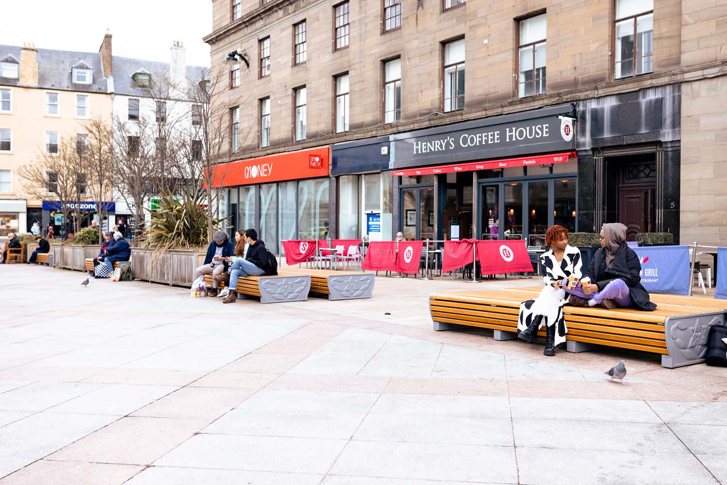 A pedestrianised street, with people sitting on benches chatting to each other. Behind them there is a sandstone building with a café on the ground floor.