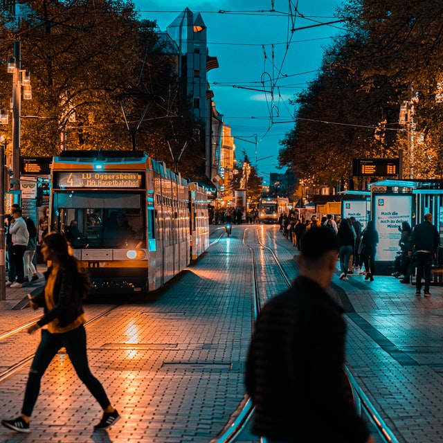 A photograph of an urban street at night - in Mannheim Germany - people cross the road in front of a tram. 