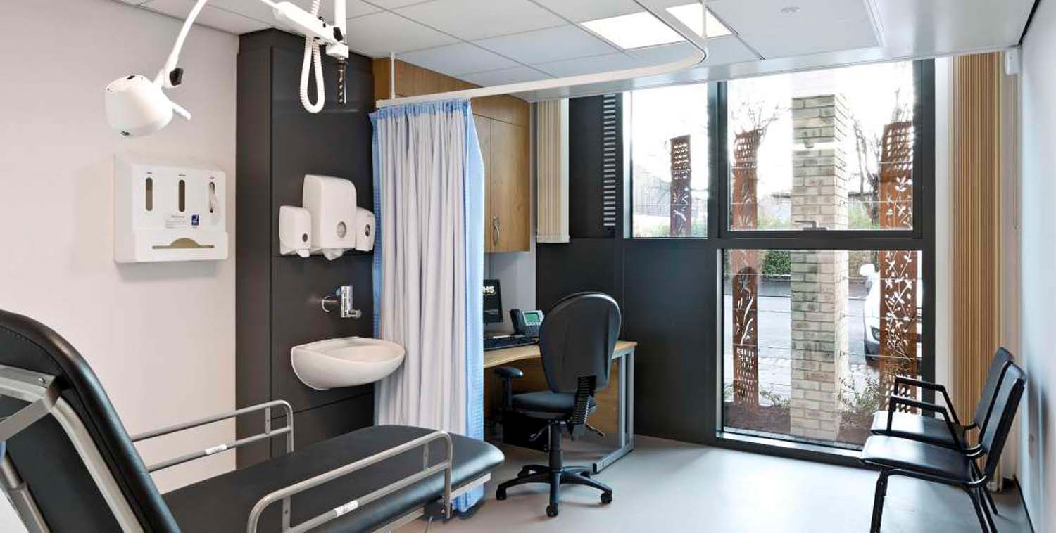 The interior of a GP consultation room with a black examination table in the foreground, a desk and chairs and a view out through the floor to ceiling windows