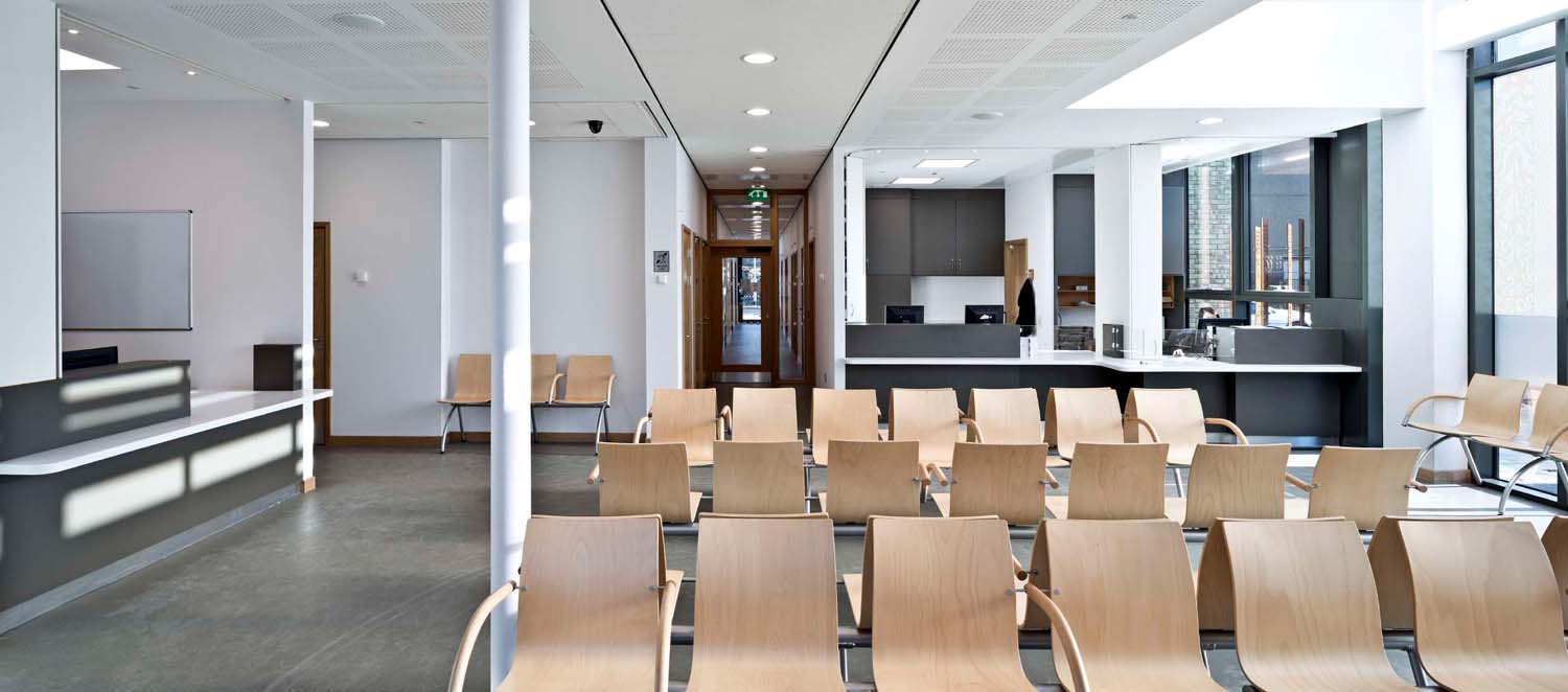 A waiting area in a health centre with black flooring, white walls and a number of rows of light coloured wooden seats