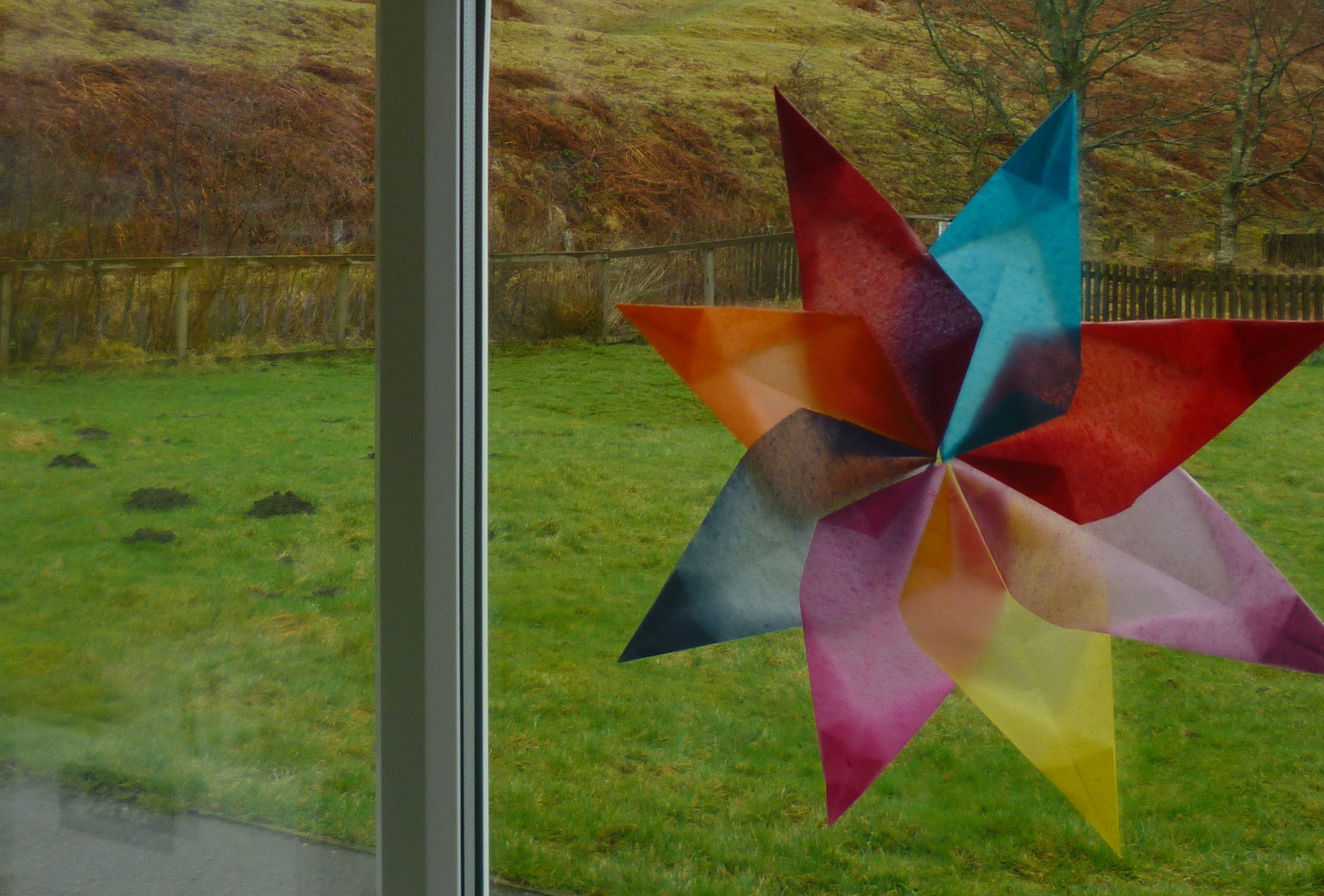 An origami of an eight point flower made out of coloured paper stuck to a window overlooking a rural landscape.