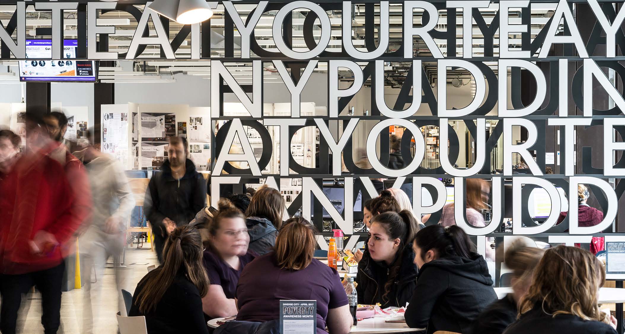 A wall sculpture in letters separates a large cafeteria in the City of Glasgow College. People are socialising, walking and eating in the space.