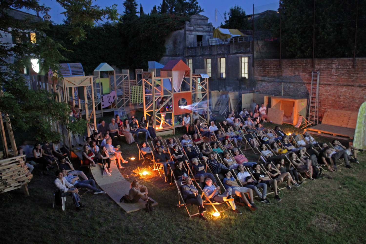 A large group of people sit in deckchairs in an enclosed garden space at night time watching a film