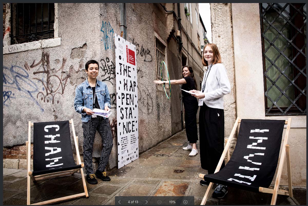 Three young people stand at the entrance to a lane holding leaflets, in front of signage and next to two black deckchairs with the words change and invite