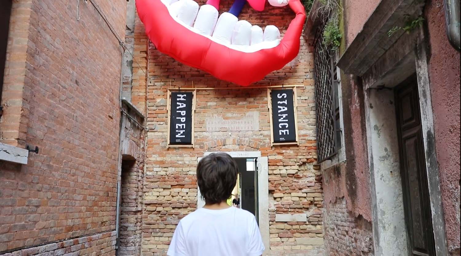 A child wearing a white t-shirt stands in a narrow streets surrounded by brick buildings looking up at a large inflatable red mouth and two black deck chairs with the words Happen and Stance 