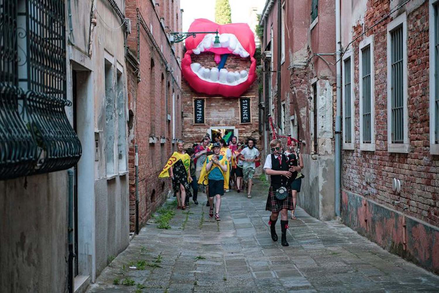 A group of people walk behind a young man playing the bagpipes down a narrow lane of stone buildings with a large inflatable smiling mouth visible in the background