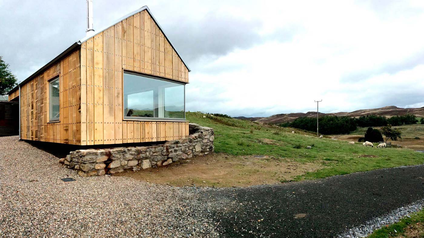 A photograph of a small timber clad building sat in a Scottish landscape, the building has a large corner window.