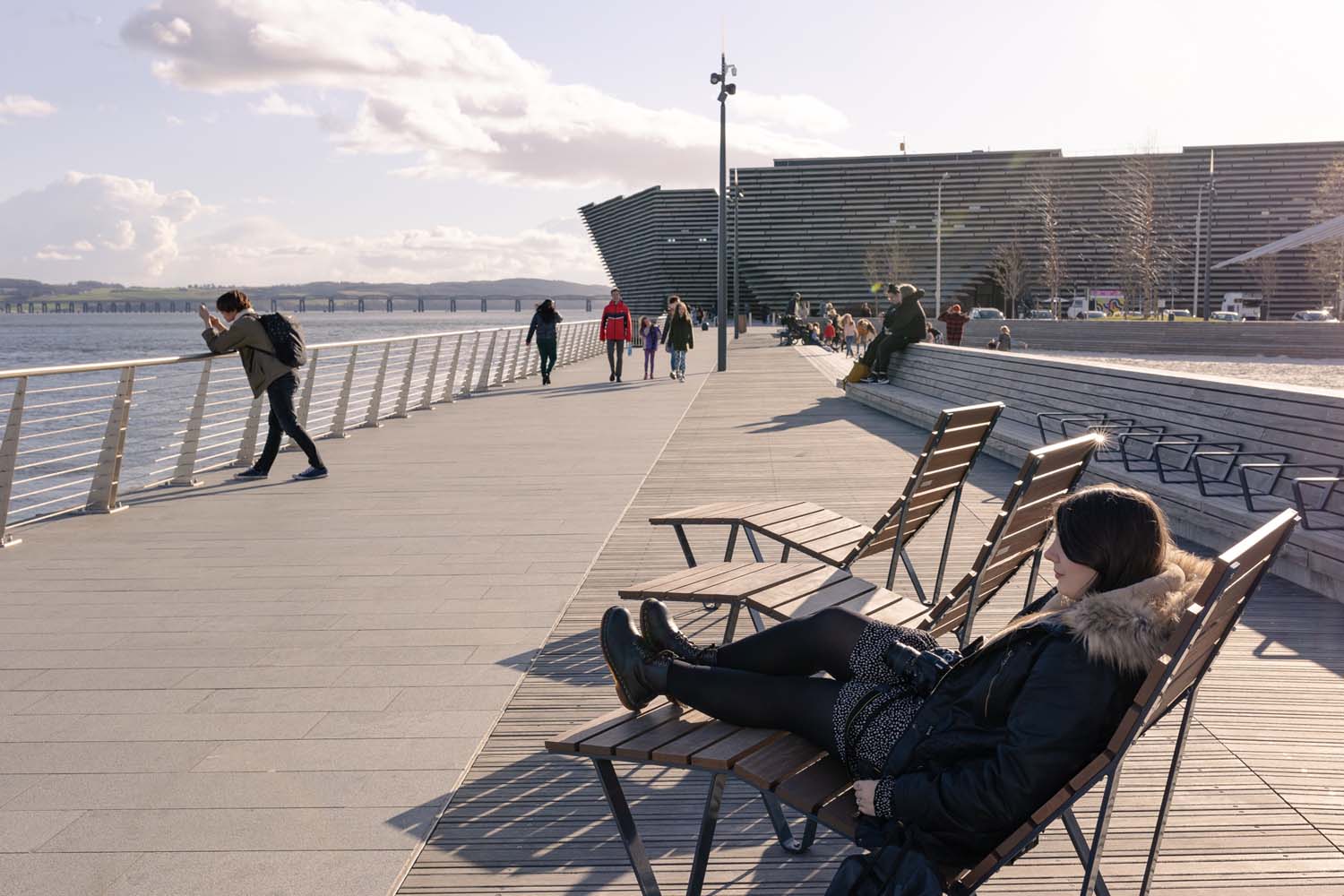 A photograph of the river front in Dundee with V&A Dundee in the background and a woman relaxing on a lounger in the sunshine, and a man looking across the Tay