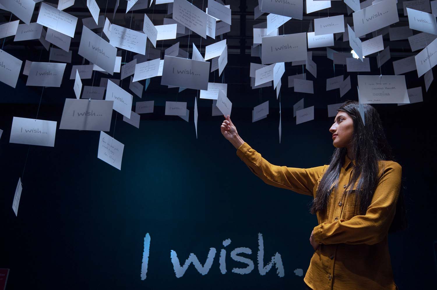 A girl in a yellow shirt looks at white postcards hanging from the ceiling in a dark room. The phrase "I wish" is written in large white letters on the wall behind her.