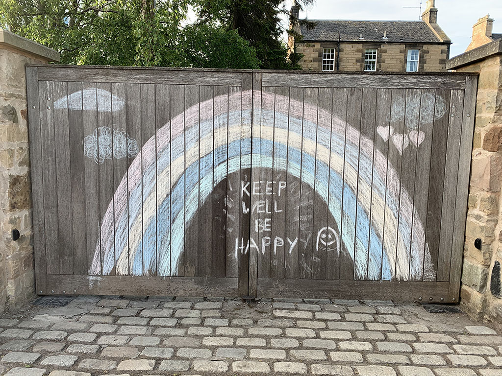 A drawing of a rainbow with chalk on a wooden gate with the words 'Keep well be happy'.