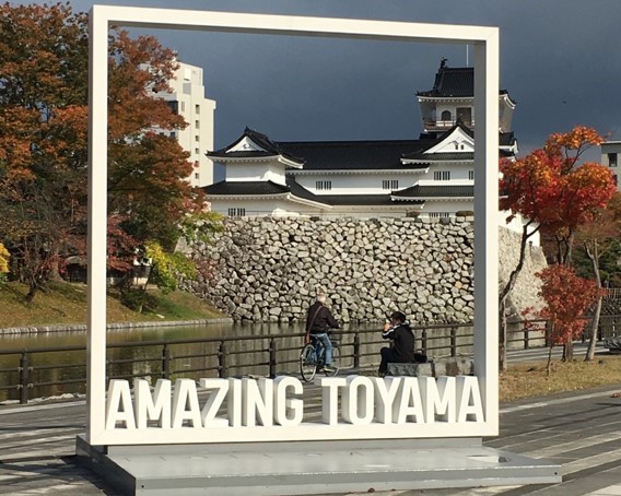 A public art sculpture consisting of the words 'Amazing Toyama' and a large white square that frames the white traditional Toyama Castle, a reconstructed flatland style castle flanked by a moat.