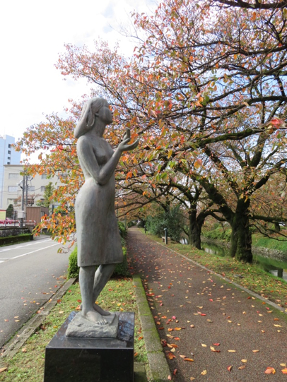 An autumnal scene as a sculpture of a woman in a skirt overlooks a bike lane and stream in a Pocket Park in Toyama, Japan