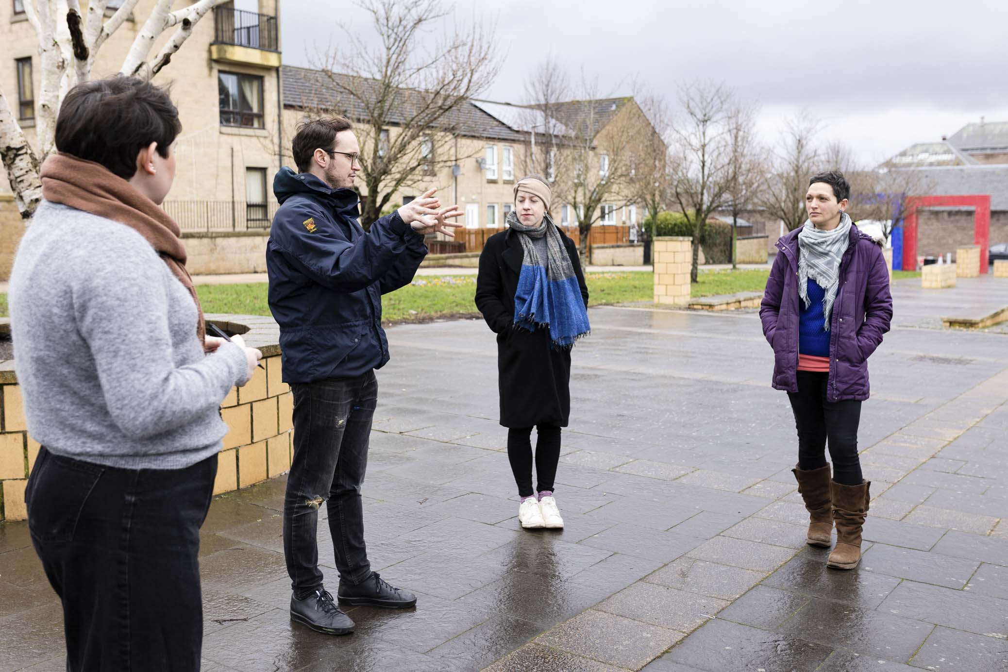 Four people wearing winter clothing stand in a circle and talk to each other at an outdoor space.