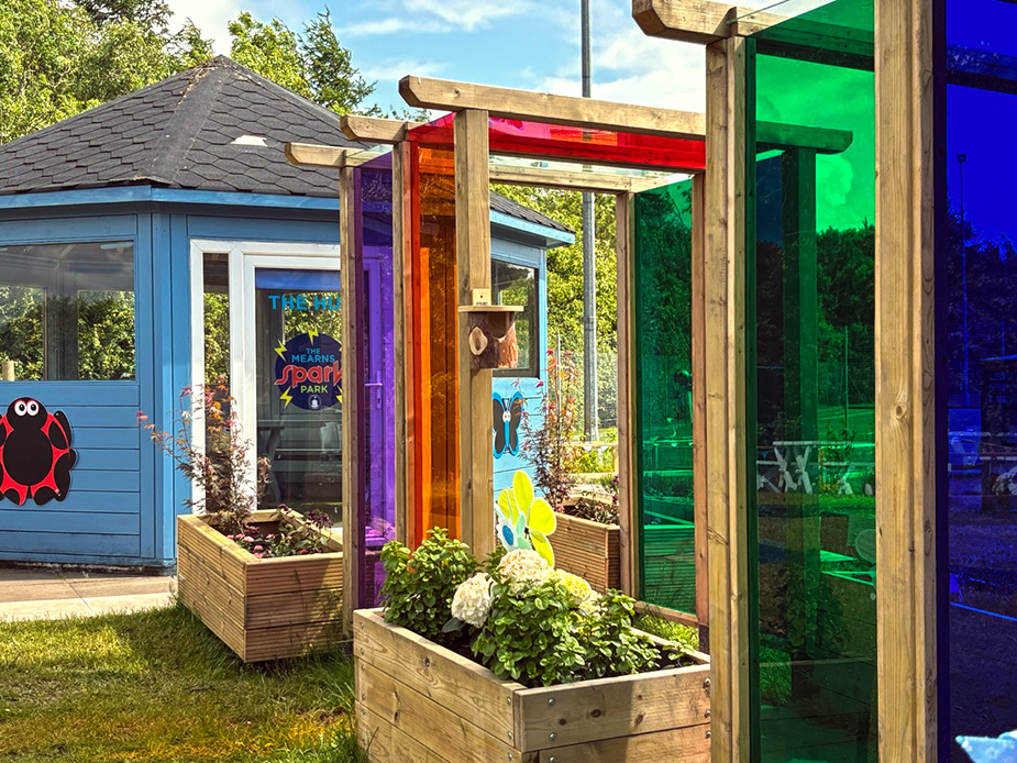 Colorful glass panels in wooden frames surround vibrant flower beds leading to a blue hut on a school ground.