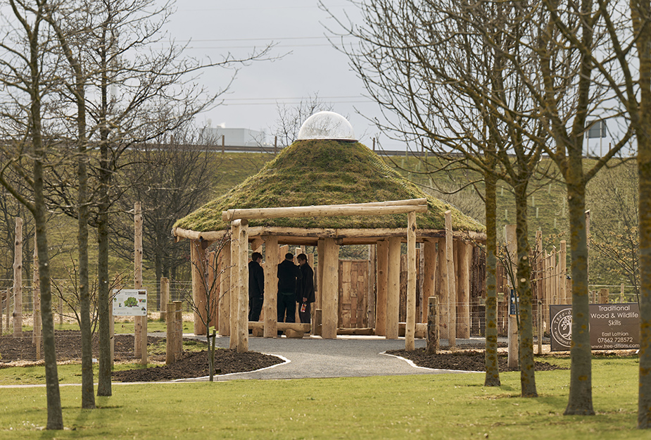 Wooden shelter with a grass-covered roof in a park, surrounded by young trees and a few people standing inside.