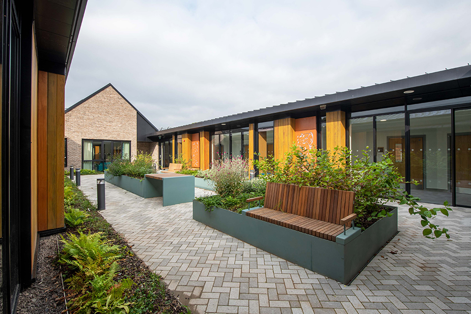 Modern hospital courtyard with wooden benches, raised flower beds, paved walkway, and greenery under a cloudy sky.
